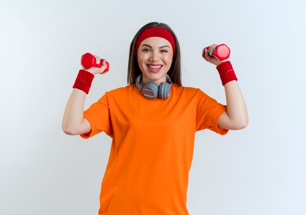 Joyful young sporty woman wearing headband and wristbands and headphones on neck raising up dumbbells isolated on white wall