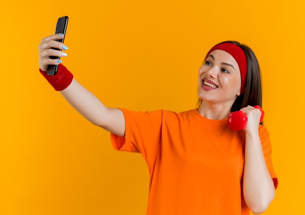 Joyful young sporty woman wearing headband and wristbands holding dumbbell and taking selfie 