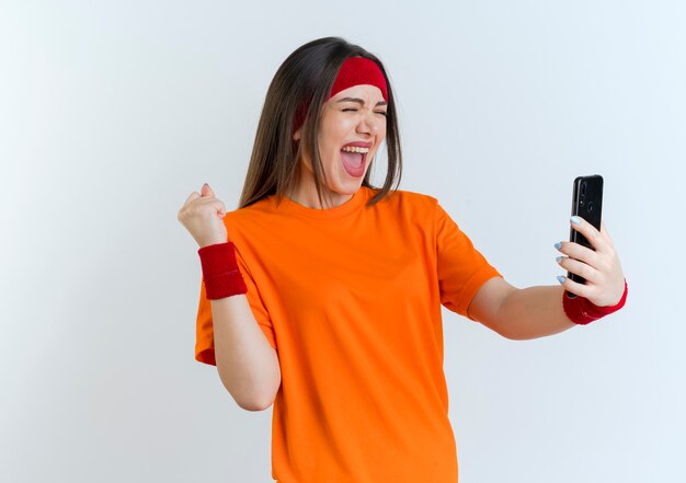 Joyful young sporty woman wearing headband and wristbands holding mobile phone doing yes gesture with closed eyes isolated on white wall with copy space