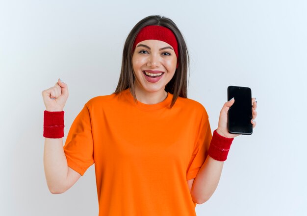 Радостная молодая спортивная женщина с повязкой на голову и браслетами, держащая мобильный телефон, смотрит, делает жест да