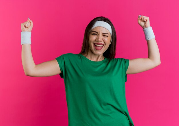 Joyful young sporty woman wearing headband and wristbands looking winking doing yes gesture 