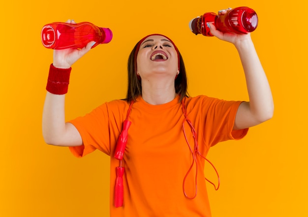 Joyful young sporty woman wearing headband and wristbands with jump rope around neck holding looking at bottles getting ready to drink water