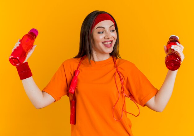 Joyful young sporty woman wearing headband and wristbands with jump rope around neck holding looking at water bottles 