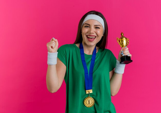 Радостная молодая спортивная женщина с повязкой на голову и браслетами с медалью на шее держит кубок победителя и делает жест
