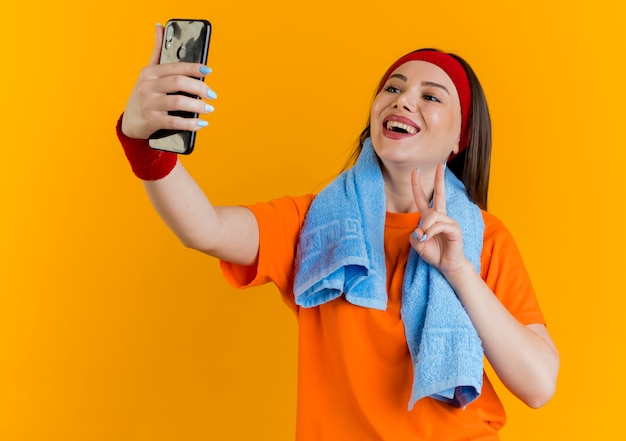 Joyful young sporty woman wearing headband and wristbands with towel around neck doing peace sign taking selfie 