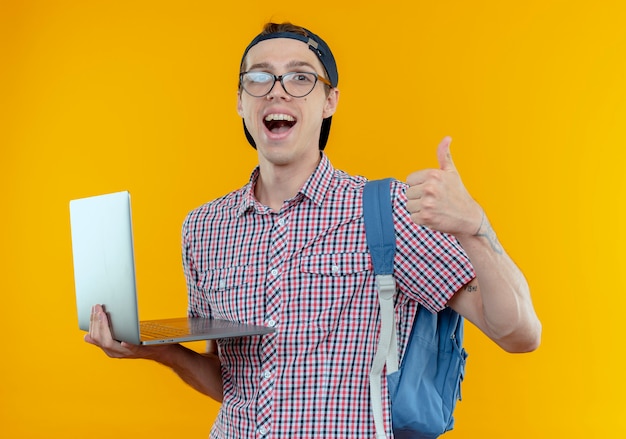 joyful young student boy wearing back bag and glasses and cap holding laptop his thumb up