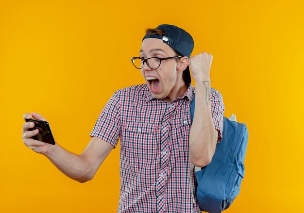 バックパックと眼鏡と帽子をかぶって携帯電話を見てはいジェスチャーを示すうれしそうな若い学生の男の子
