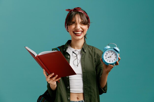 Joyful young student girl wearing bandana and backpack holding open note pad showing alarm clock with closed eyes putting glasses on her blouse isolated on blue background