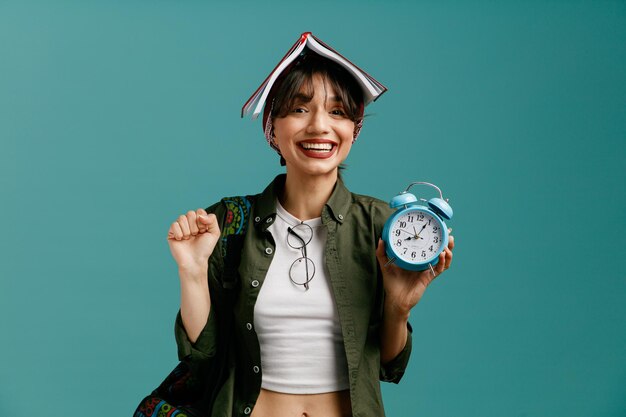 joyful young student girl wearing bandana and backpack putting glasses on her blouse holding open note pad on her head looking at camera showing alarm clock and yes gesture isolated on blue background