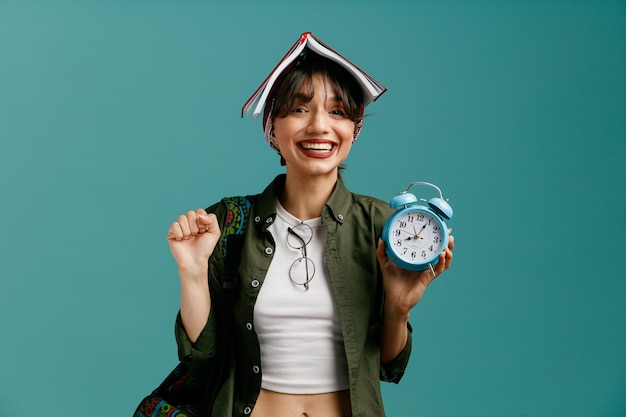 joyful young student girl wearing bandana and backpack putting glasses on her blouse holding open note pad on her head looking at camera showing alarm clock and yes gesture isolated on blue background