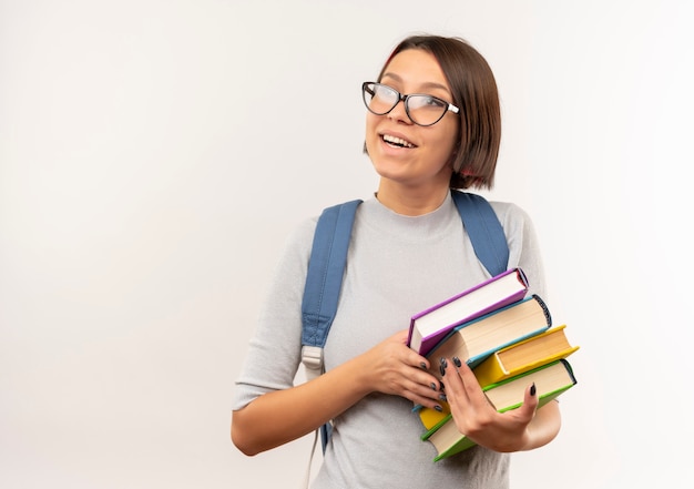 Joyful young student girl wearing glasses and back bag holding books looking at side isolated on white