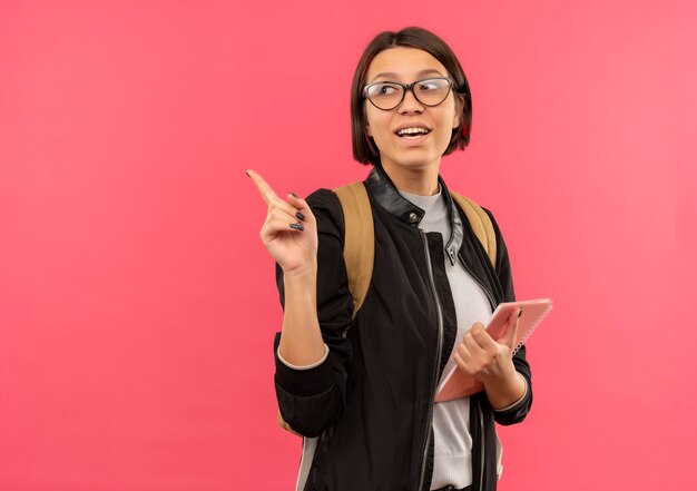 Joyful young student girl wearing glasses and back bag holding note pad pointing and looking at side isolated on pink