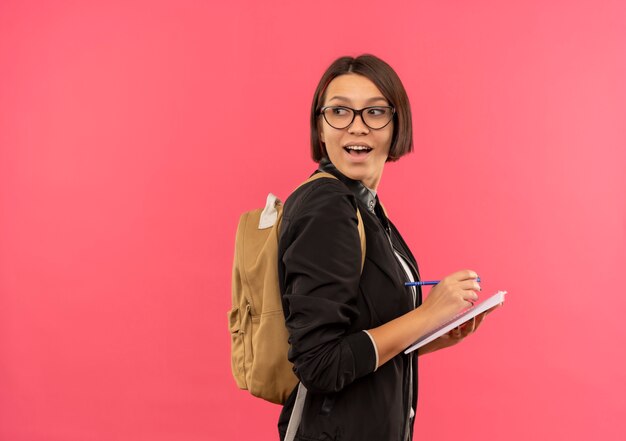 Joyful young student girl wearing glasses and back bag standing in profile view holding pen and note pad looking behind isolated on pink