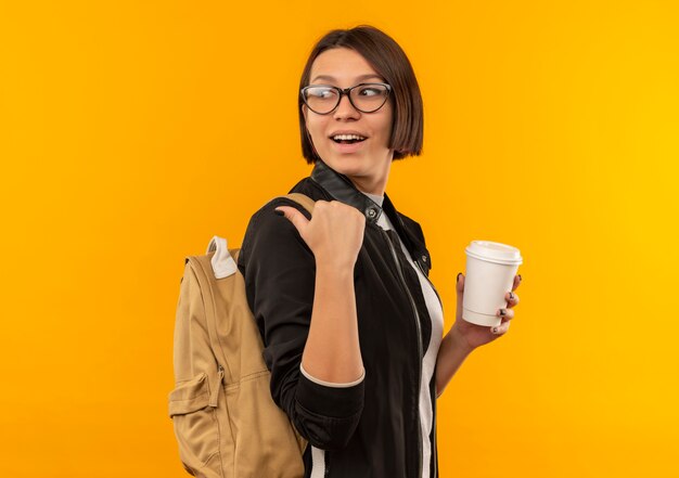 Joyful young student girl wearing glasses and back bag standing in profile view holding plastic coffee cup pointing and looking behind isolated on orange