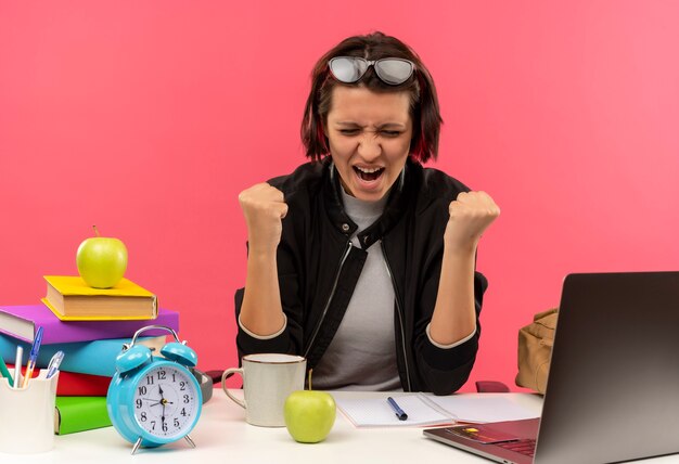 Joyful young student girl wearing glasses on head sitting at desk doing homework clenching fists with closed eyes isolated on pink