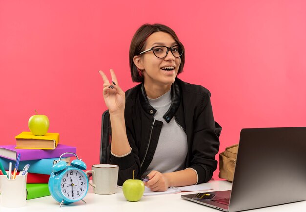 Joyful young student girl wearing glasses sitting at desk doing peace sign isolated on pink