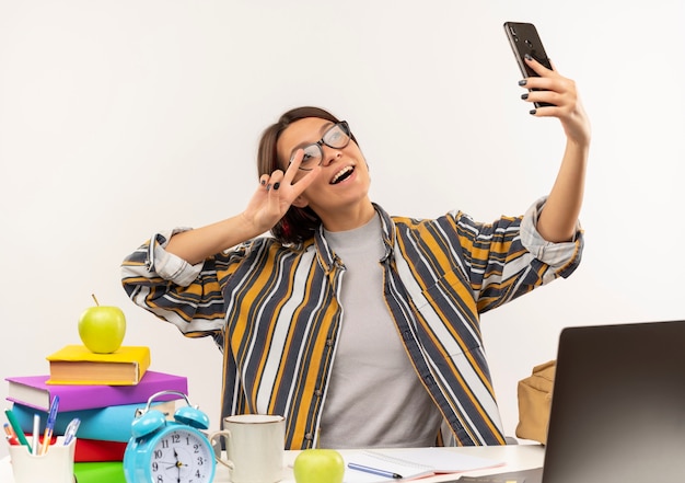 Gioiosa ragazza giovane studente con gli occhiali seduto alla scrivania facendo segno di pace tenendo selfie con il telefono cellulare isolato su bianco