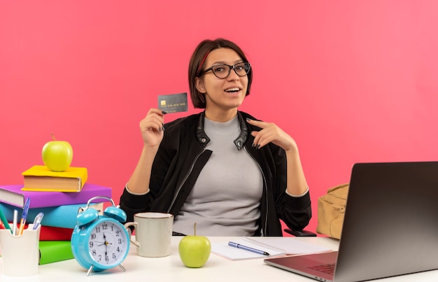 Joyful young student girl wearing glasses sitting at desk holding and pointing at credit card doing homework isolated on pink