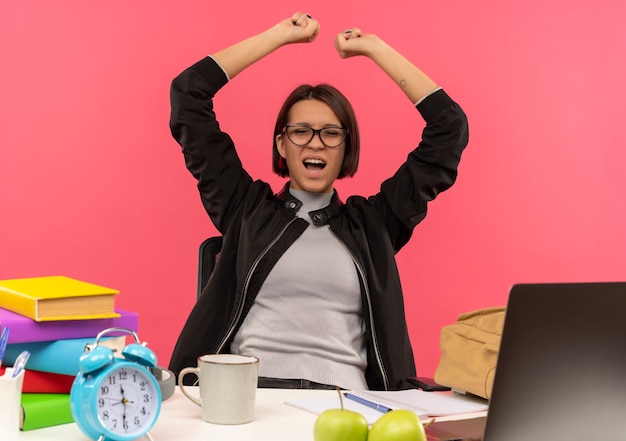 Joyful young student girl wearing glasses sitting at desk raising fists with closed eyes doing homework isolated on pink