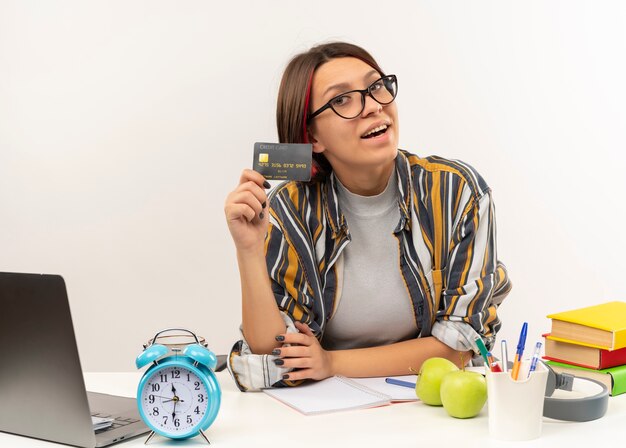 Joyful young student girl wearing glasses sitting at desk showing credit card isolated on white