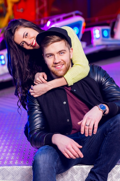 Joyful young stylish  couple being playful while visiting an attractions park arcade with rides