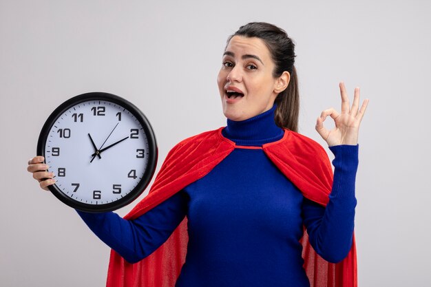 Joyful young superhero girl holding wall clock and showing okay gesture isolated on white