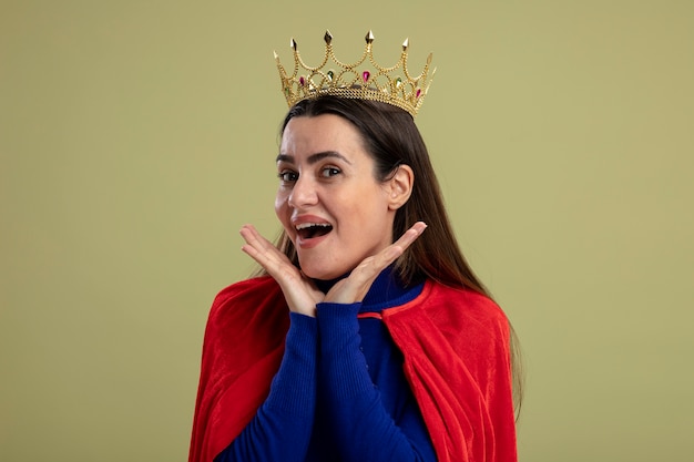 Free photo joyful young superhero girl wearing crown putting hands on cheeks isolated on olive green background