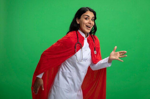 Радостная молодая девушка супергероя в медицинском халате со стетоскопом, показывающая стиль робота, изолированного на зеленом
