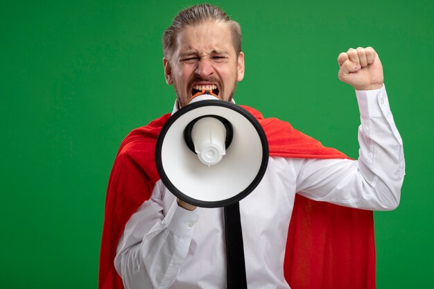 Joyful young superhero guy speaks on loudspeakes and raising fist isolated on green background