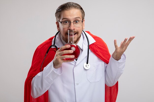 Joyful young superhero guy wearing medical robe with stethoscope and glasses holding chemistry glass bottle