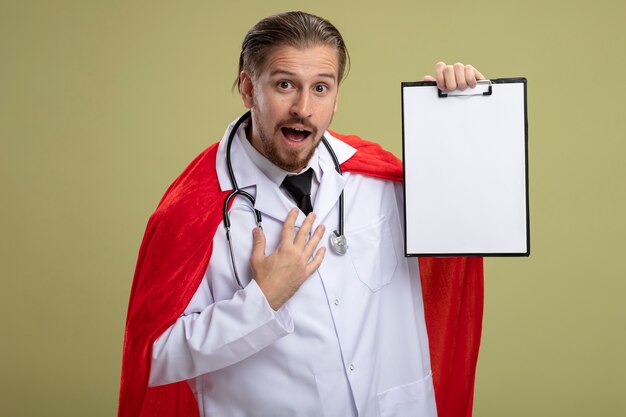 Joyful young superhero guy wearing stethoscope with medical robe holding clipboard and putting hand on chest isolated on olive green background