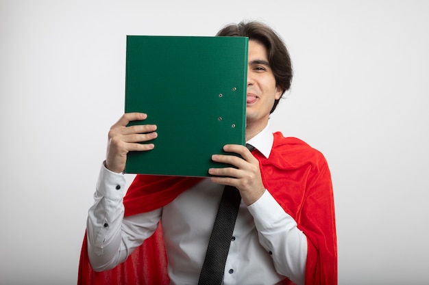 Joyful young superhero guy wearing tie covered face with clipboard and showing tongue isolated on white background