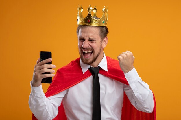 Радостный молодой супергерой в галстуке и короне, держащий и смотрящий в телефон, показывая жест да, изолированный на оранжевом фоне