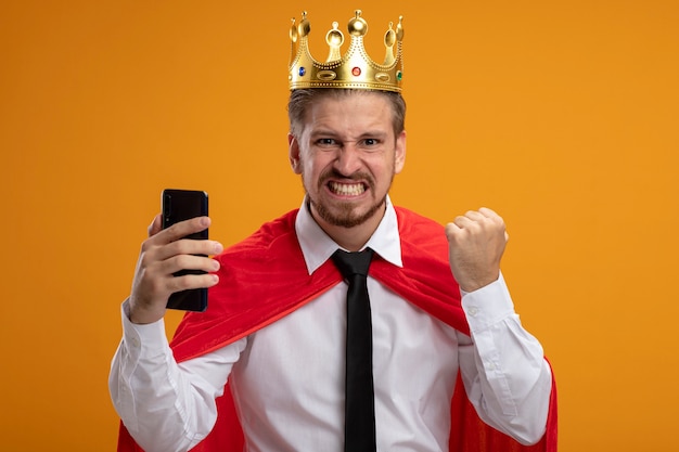 Joyful young superhero guy wearing tie and crown holding phone and showing yes gesture isolated on orange background