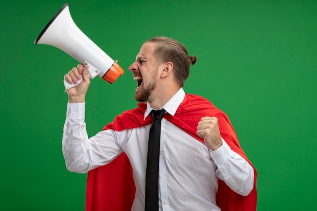 Free photo joyful young superhero guy wearing tie speaks on loudspeaker and showing yes gesture isolated on green