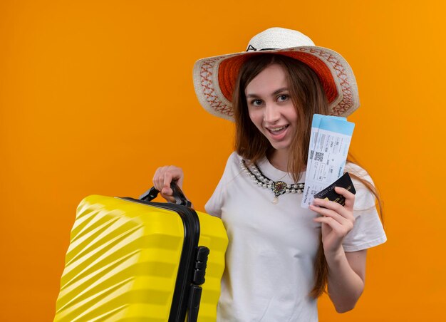 격리 된 오렌지 공간에 가방과 비행기 티켓, 신용 카드를 들고 모자를 쓰고 즐거운 젊은 여행자 소녀