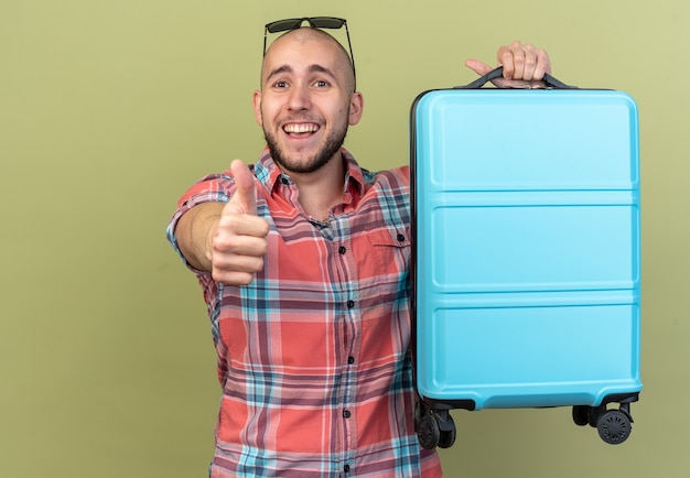 Joyful young traveler man holding suitcase and thumbing up isolated on olive green wall with copy space