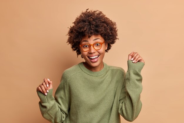 Joyful young woman dances over beige wall keeps hands up moves with rhythm of music being on party expreses positive emotions isolated over brown wall