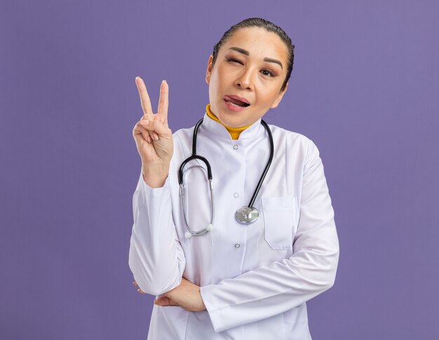 joyful young woman doctor in white medicine coat with stethoscope around neck showing v-sign sticking out tongue winking happy and positive standing over purple wall