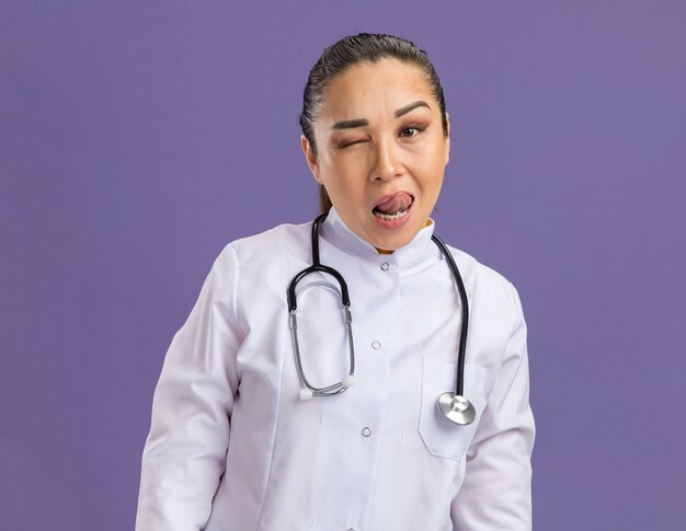 Joyful young woman doctor in white medicine coat with stethoscope around neck  winking sticking out tongue standing over purple wall