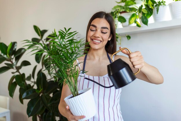 うれしそうな若い女性は家で彼女の時間を楽しんで、家の窓のそばで彼女の植物に水をまく女性は家の庭でリラックスした日に木の上の彼女のシダの水を世話します