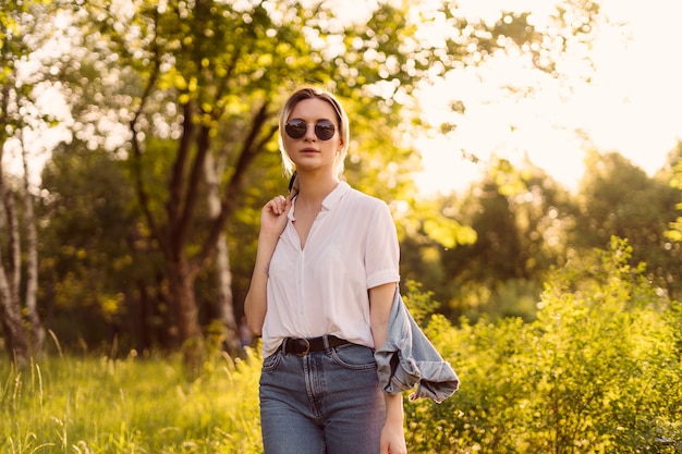 Gioiosa giovane donna con occhiali da sole alla moda che cammina nella natura in una bella giornata