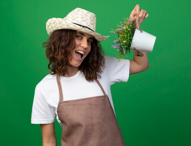 Радостная молодая женщина-садовник в униформе в садовой шляпе держит цветок в цветочном горшке, изолированном на зеленом
