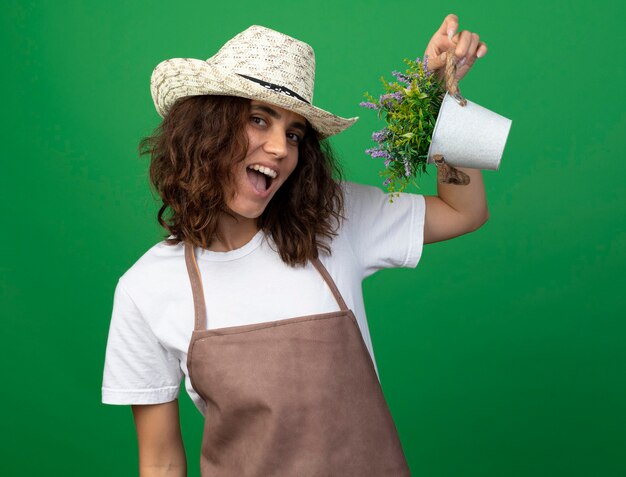 緑に分離された植木鉢の花を保持しているガーデニング帽子を身に着けている制服を着たうれしそうな若い女性の庭師