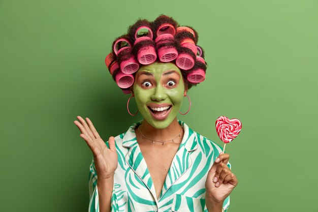 joyful young woman keeps hand raised, holds lollipop, smiles broadly, applies beauty mask to look young, makes hairdo with hair rollers, isolated on green wall. Hairstyling, face care