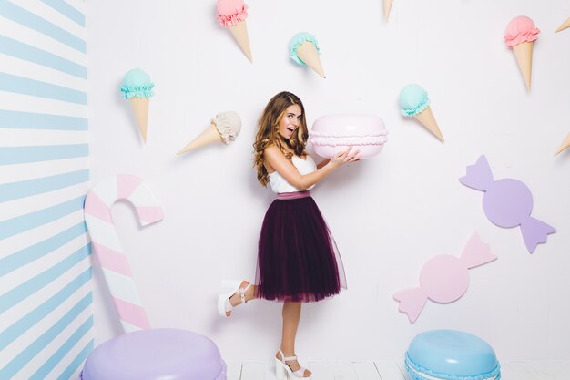 Joyful young woman in tulle skirt having fun with big macaron among sweets. Pastel colors, ice cream, happiness, cupcakes, smiling, surprised, playful.
