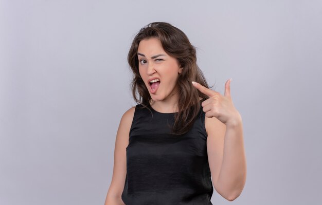 Joyful young woman wearing black undershirt blinked and point to side on white wall