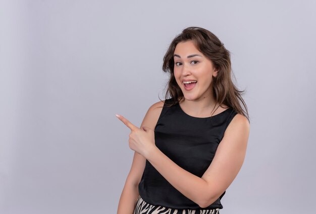 Joyful young woman wearing black undershirt points to side on white wall