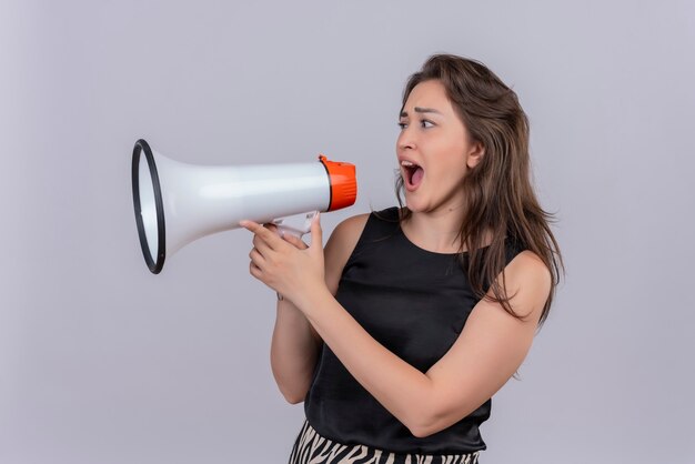 Joyful young woman wearing black undershirt speaks through loudspeakers on white wall