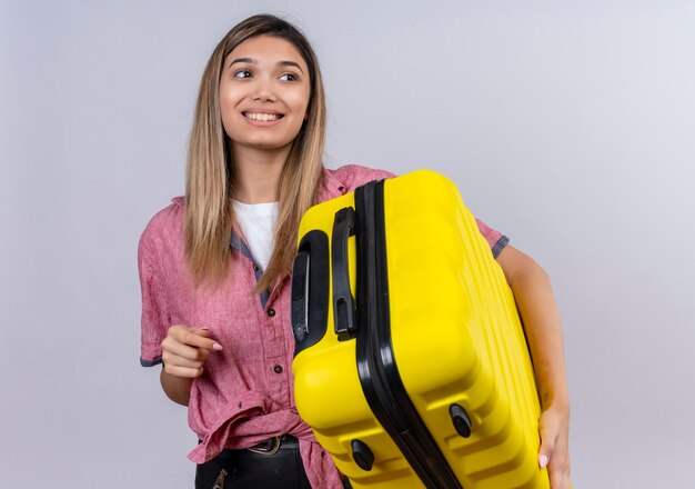 Радостная молодая женщина в красной рубашке держит желтый чемодан, глядя в сторону на белой стене